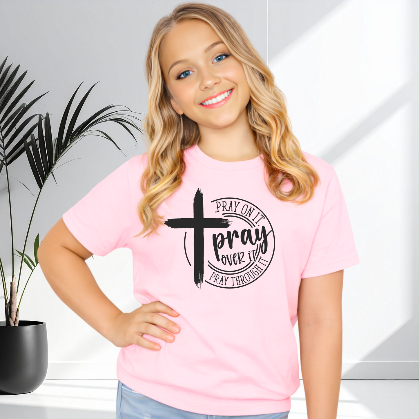 Young girl wearing a pink t-shirt with a cross and text design, standing against a light background.