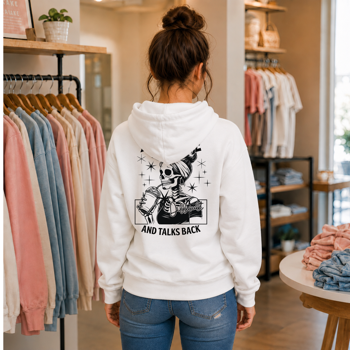 Woman showing the back of a white hoodie with a black Expensive and Talks Back skeleton-woman graphic in a boutique.