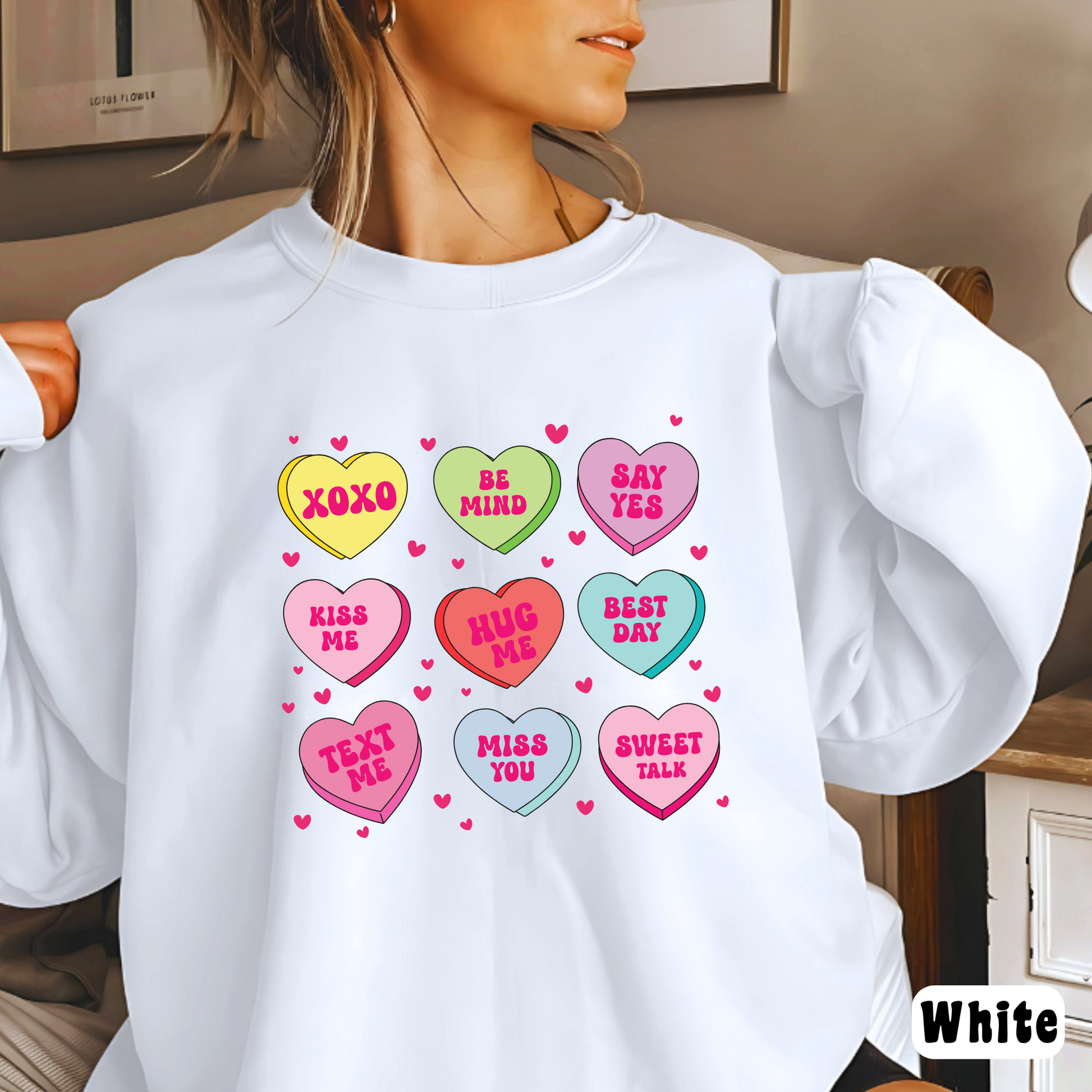 Woman wearing a white Valentine’s sweatshirt featuring colorful candy conversation hearts with messages like “XOXO,” “Be Mine,” “Say Yes,” “Kiss Me,” “Hug Me,” and more.