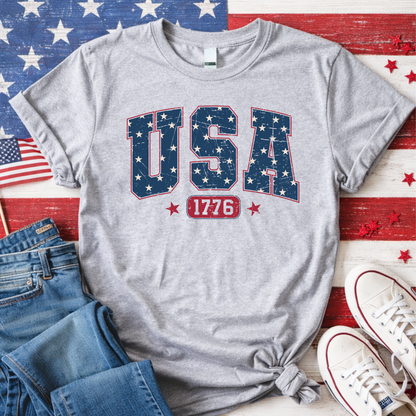 America 250 USA 1776 patriotic t-shirt flat lay on American flag background, gray tee celebrating 1776 to 2026