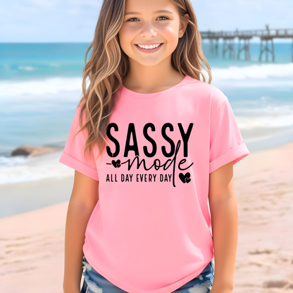 Young girl wearing a pink t-shirt with 'Sassy mode all day every day' text on a beach.