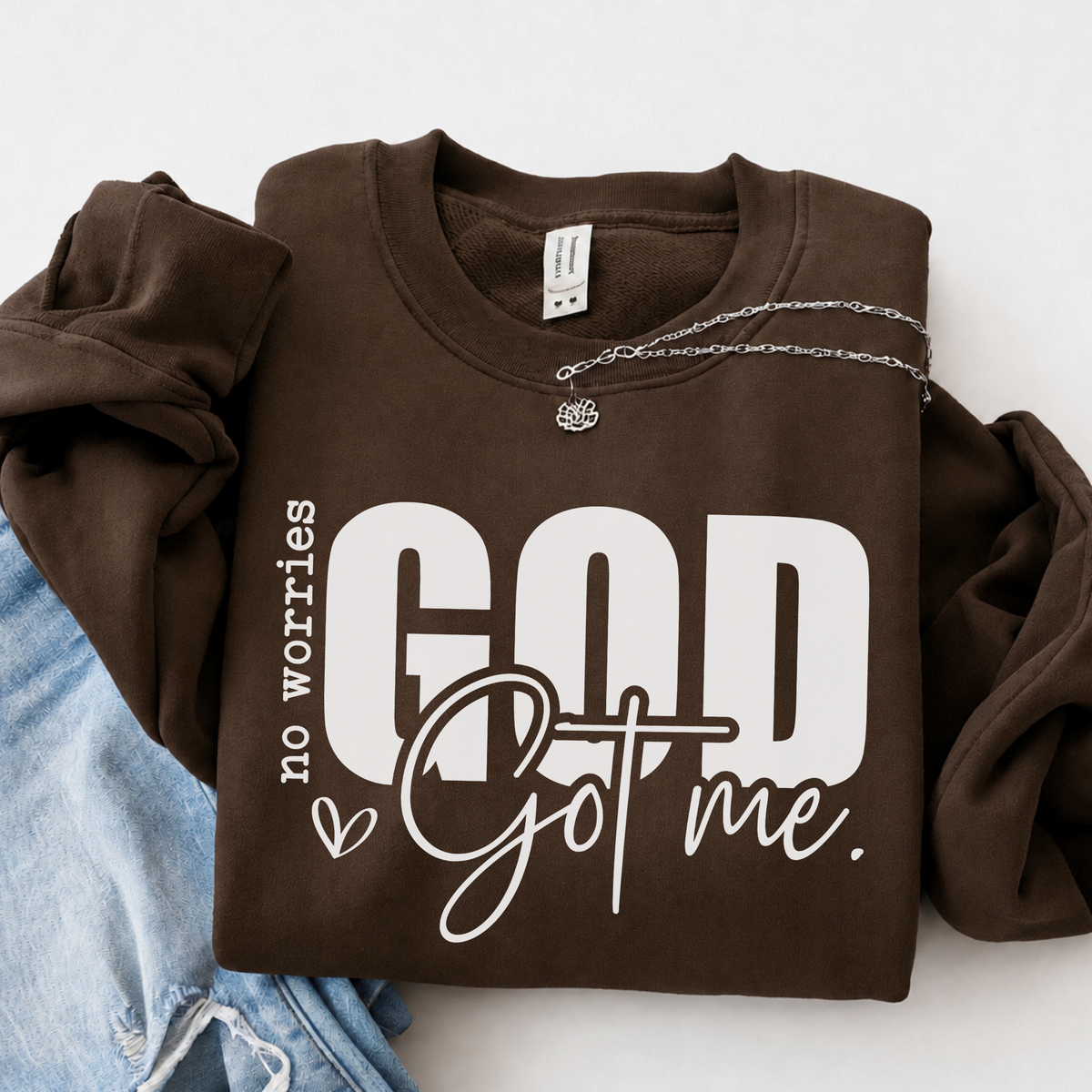 Brown No Worries God Got Me crewneck sweatshirt flat lay with bold white lettering, styled with denim.