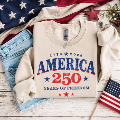 Beige t-shirt with '1776 2026 AMERICA 250 YEARS OF FREEDOM' text, American flag, and denim jeans on a wooden surface.