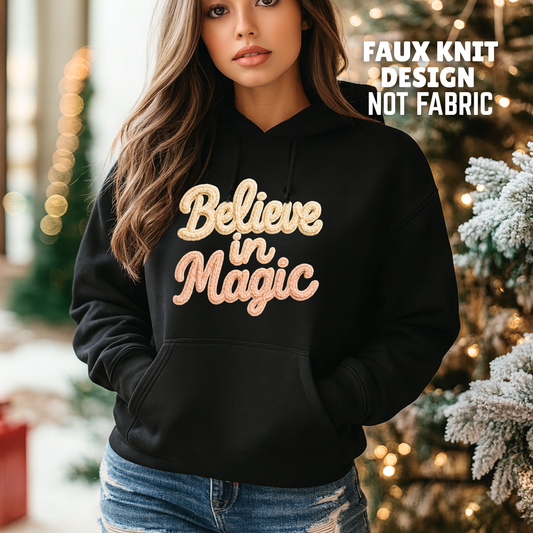 Woman wearing black hoodie with Believe in Magic faux crochet Christmas design, soft printed yarn texture, cozy winter hoodie, Black Friday holiday apparel.