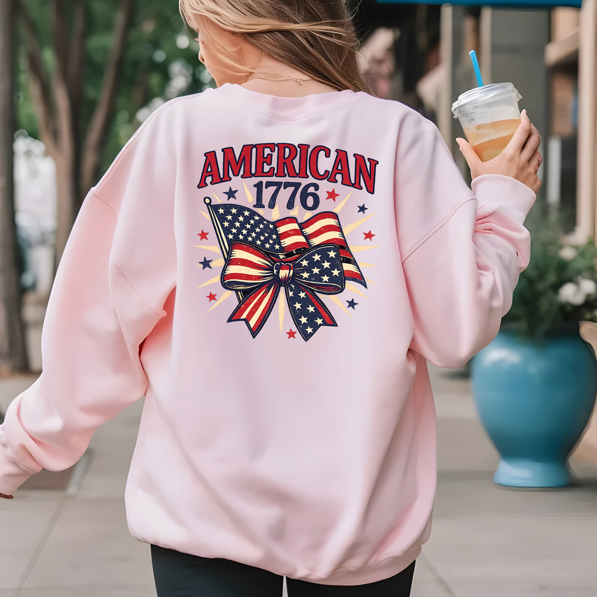 Person wearing a pink sweatshirt with 'American 1776' design, holding a drink.