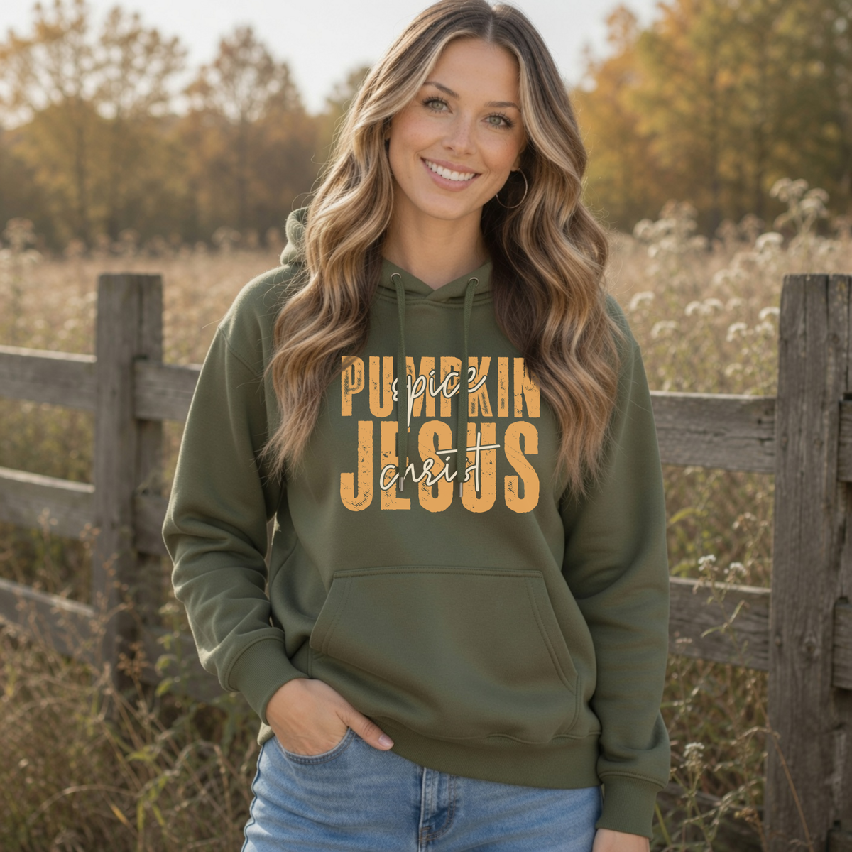 Woman in a military green Pumpkin Spice and Jesus Christ hoodie standing by a rustic fence in a fall field
