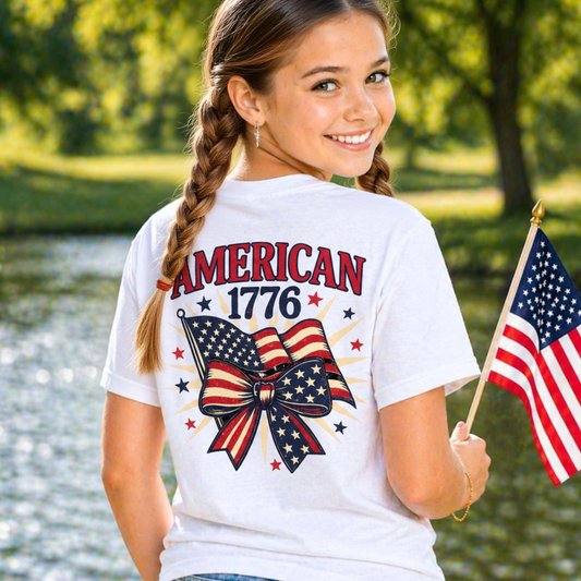 Girl wearing a white t-shirt with 'American 1776' design, holding an American flag, standing by a body of water.