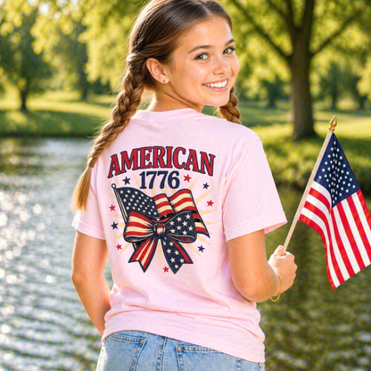 Girl wearing a pink t-shirt with 'American 1776' design, holding an American flag, standing by a body of water.