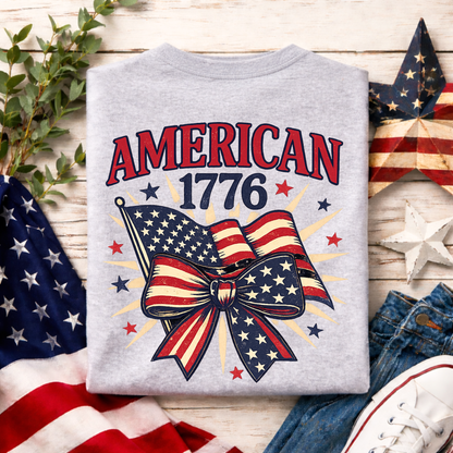 Gray sweatshirt with 'American 1776' design featuring an American flag bow, placed on a wooden surface with flags and denim jeans.