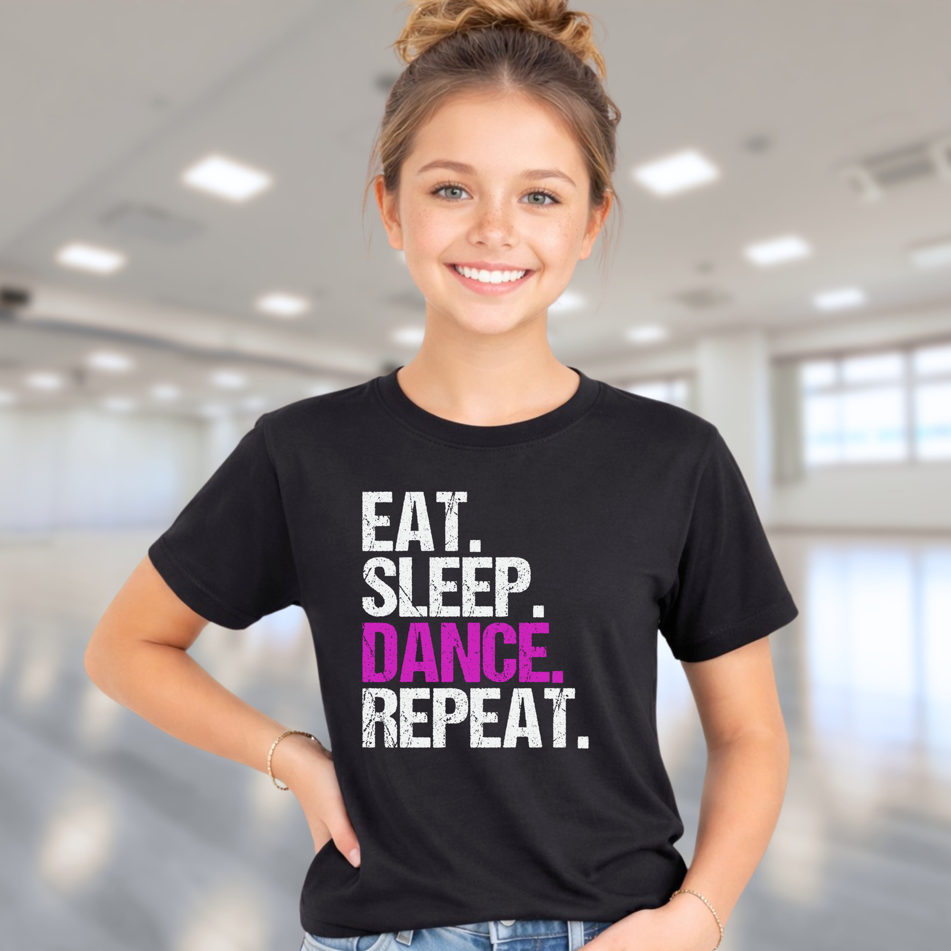 Young girl wearing a black t-shirt with 'EAT. SLEEP. DANCE. REPEAT.' text in a modern indoor setting.