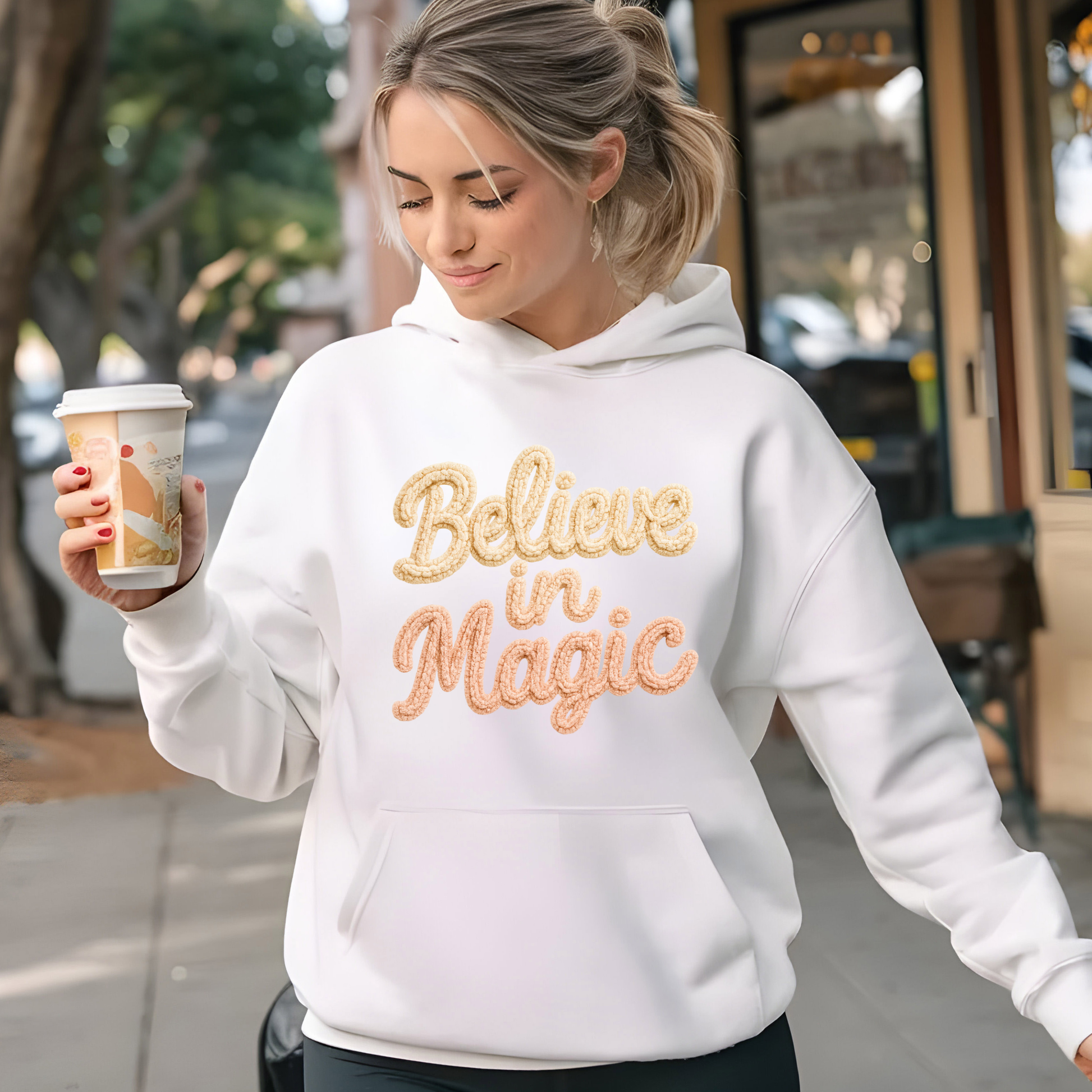 Woman wearing a white hoodie with 'Believe in Magic' text, holding a coffee cup outdoors.