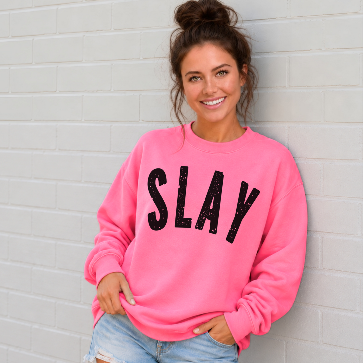 Woman wearing a bright pink SLAY crewneck sweatshirt with bold black distressed lettering against a white brick wall.