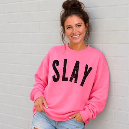 Woman wearing a bright pink SLAY crewneck sweatshirt with bold black distressed lettering against a white brick wall.