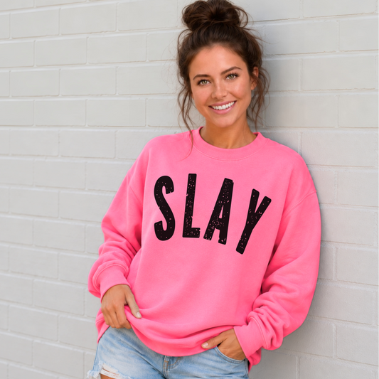 Woman wearing a bright pink SLAY crewneck sweatshirt with bold black distressed lettering against a white brick wall.