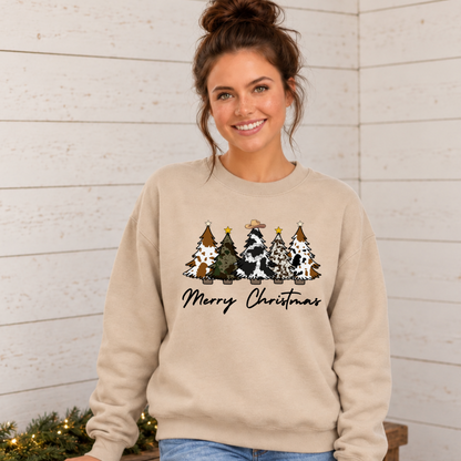 Woman wearing a sand sweatshirt with cow print Christmas trees and Merry Christmas lettering against a light shiplap wall.