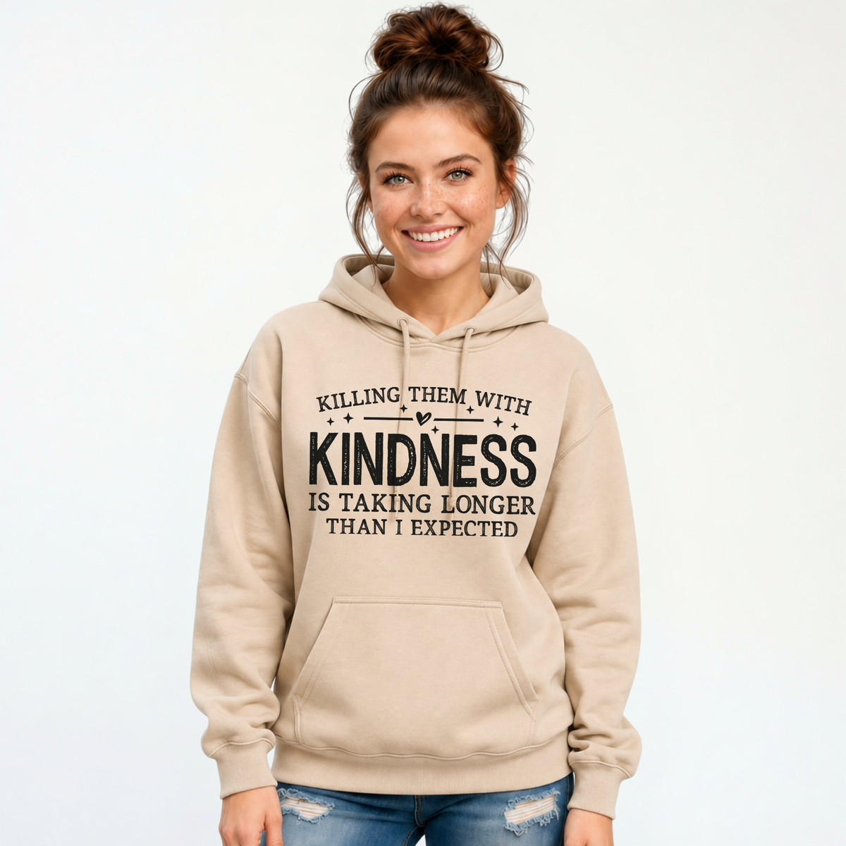 Smiling woman modeling a sand Killing Them With Kindness hoodie with distressed jeans against a white background.