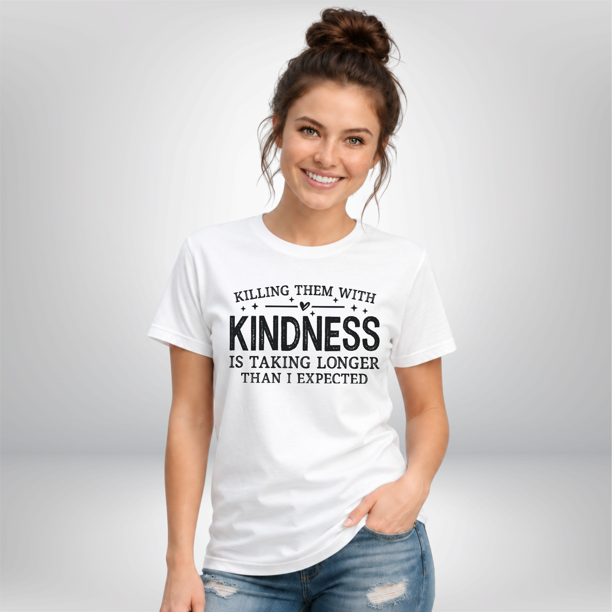 Woman against a gray background wearing a white Killing Them With Kindness T-shirt with black lettering.