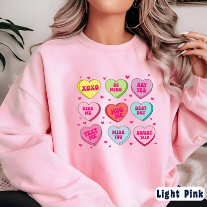 Woman wearing a light pink Valentine’s sweatshirt featuring colorful candy conversation hearts with messages like “XOXO,” “Be Mine,” “Say Yes,” “Kiss Me,” “Hug Me,” and more.