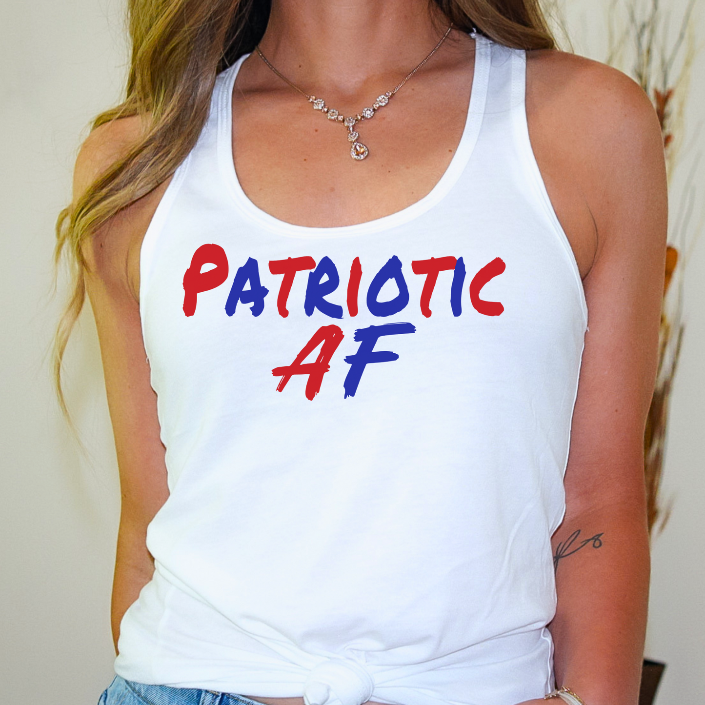 White racerback tank top tied at waist modeled indoors with "Patriotic AF" design.

