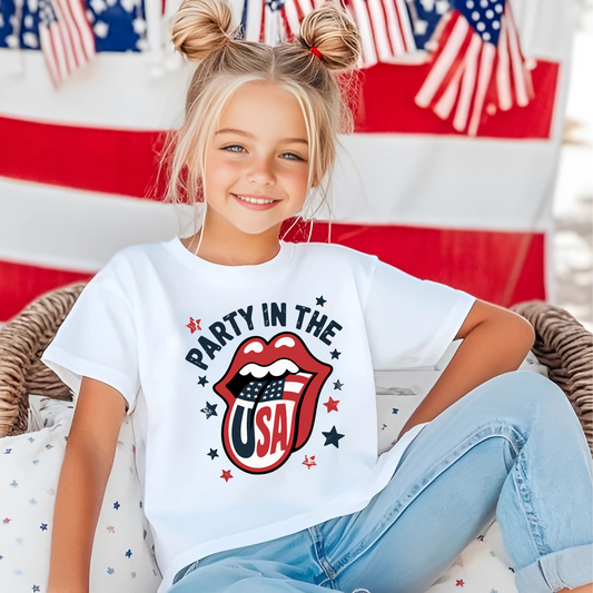 Blonde girl in space buns wearing white “Party in the USA” tee with patriotic lips design in front of American flag backdrop. Cute and festive kids’ shirt for Independence Day, USA parades, fireworks, and summer celebrations.

