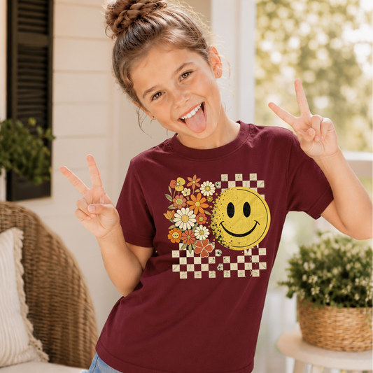 Young girl wearing a maroon t-shirt with a smiley face and floral design, making peace signs.