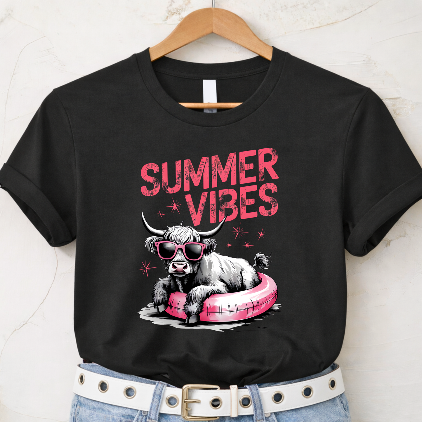 Black t-shirt with a graphic of a highland cow on an inner tube and 'Summer Vibes' text, hanging on a wooden hanger.