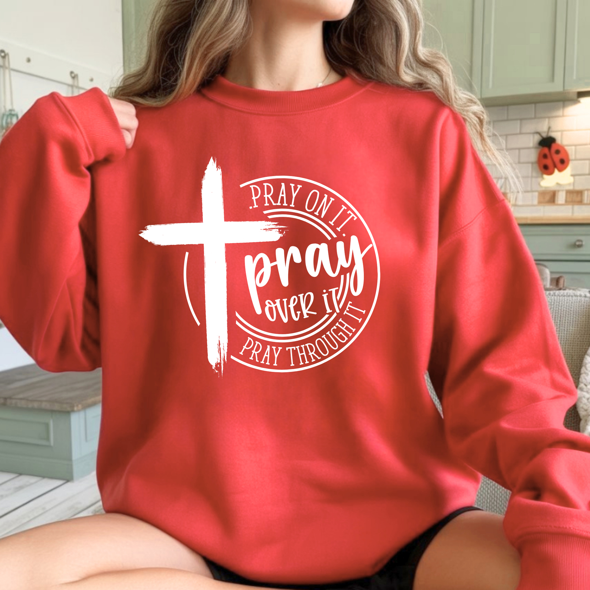 Person wearing a red sweatshirt with a white cross and text design in a kitchen setting.
