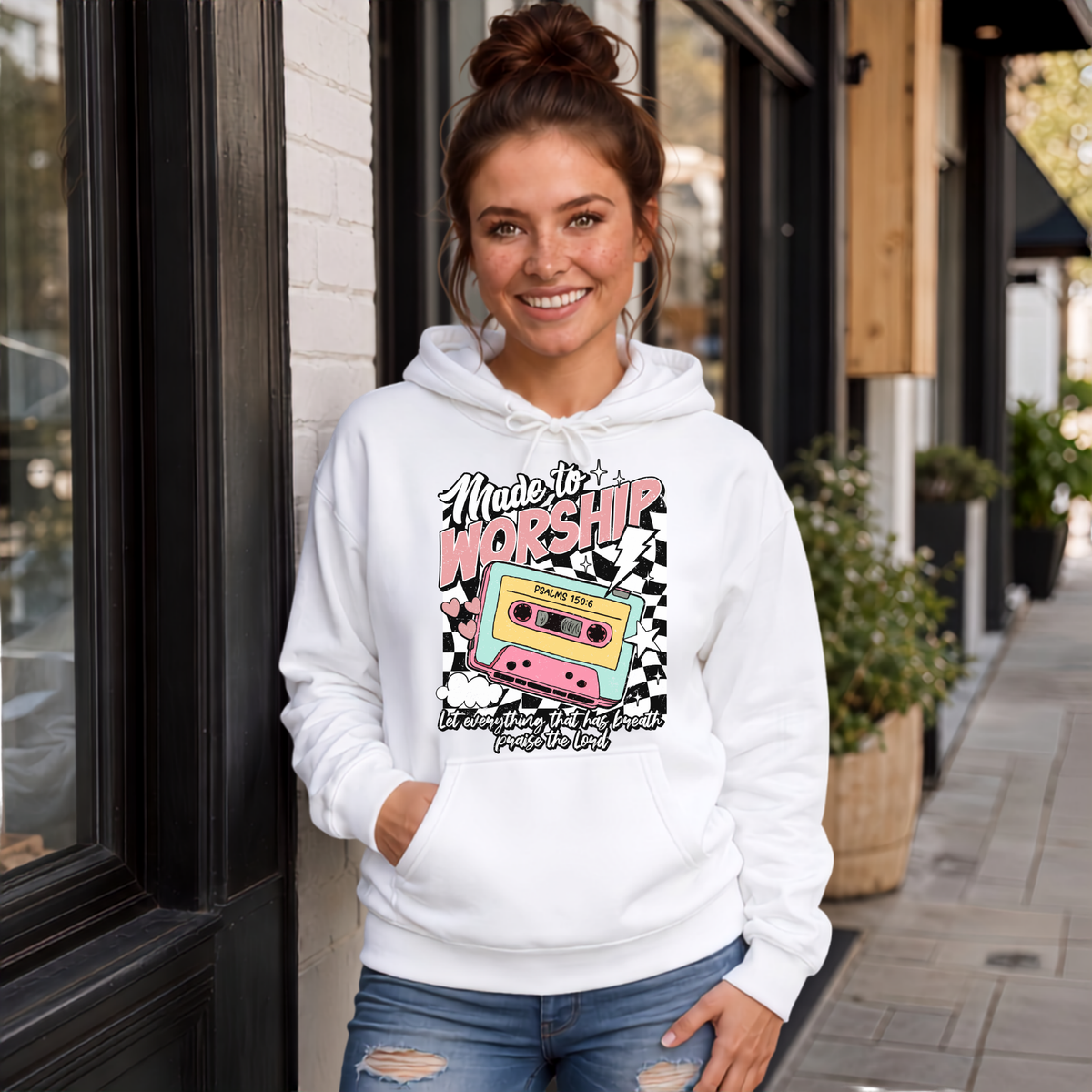 Woman wearing a white Made to Worship hoodie with a retro cassette graphic outside a storefront.