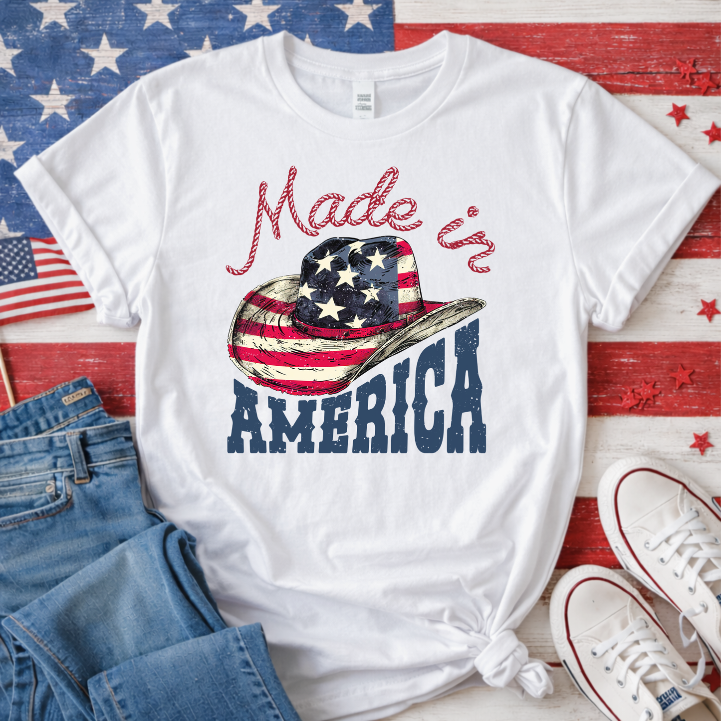 White t-shirt with 'Made in America' graphic on a wooden surface with American flags and jeans.