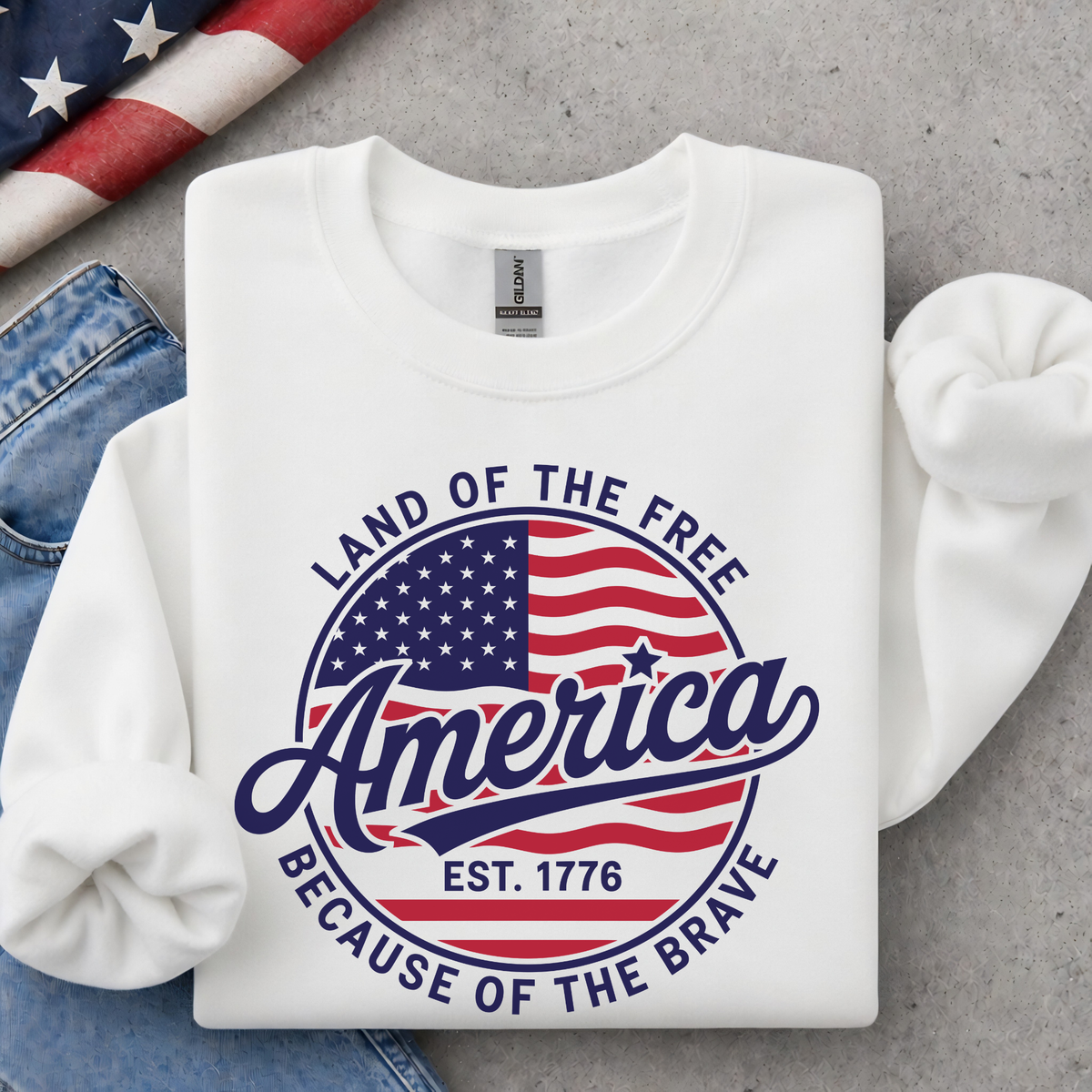 Folded white crewneck sweatshirt with a red, white, and blue circular America graphic reading Land of the Free, Est. 1776, and Because of the Brave, styled with jeans and an American flag.