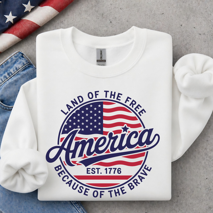 Folded white crewneck sweatshirt with a red, white, and blue circular America graphic reading Land of the Free, Est. 1776, and Because of the Brave, styled with jeans and an American flag.