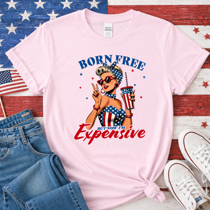 Pink t-shirt with graphic design featuring a woman in an American flag bikini, sunglasses, and a cup of ice cream. Text reads 'Born Free But Now I'm Expensive' on a wooden surface with an American flag in the background.
