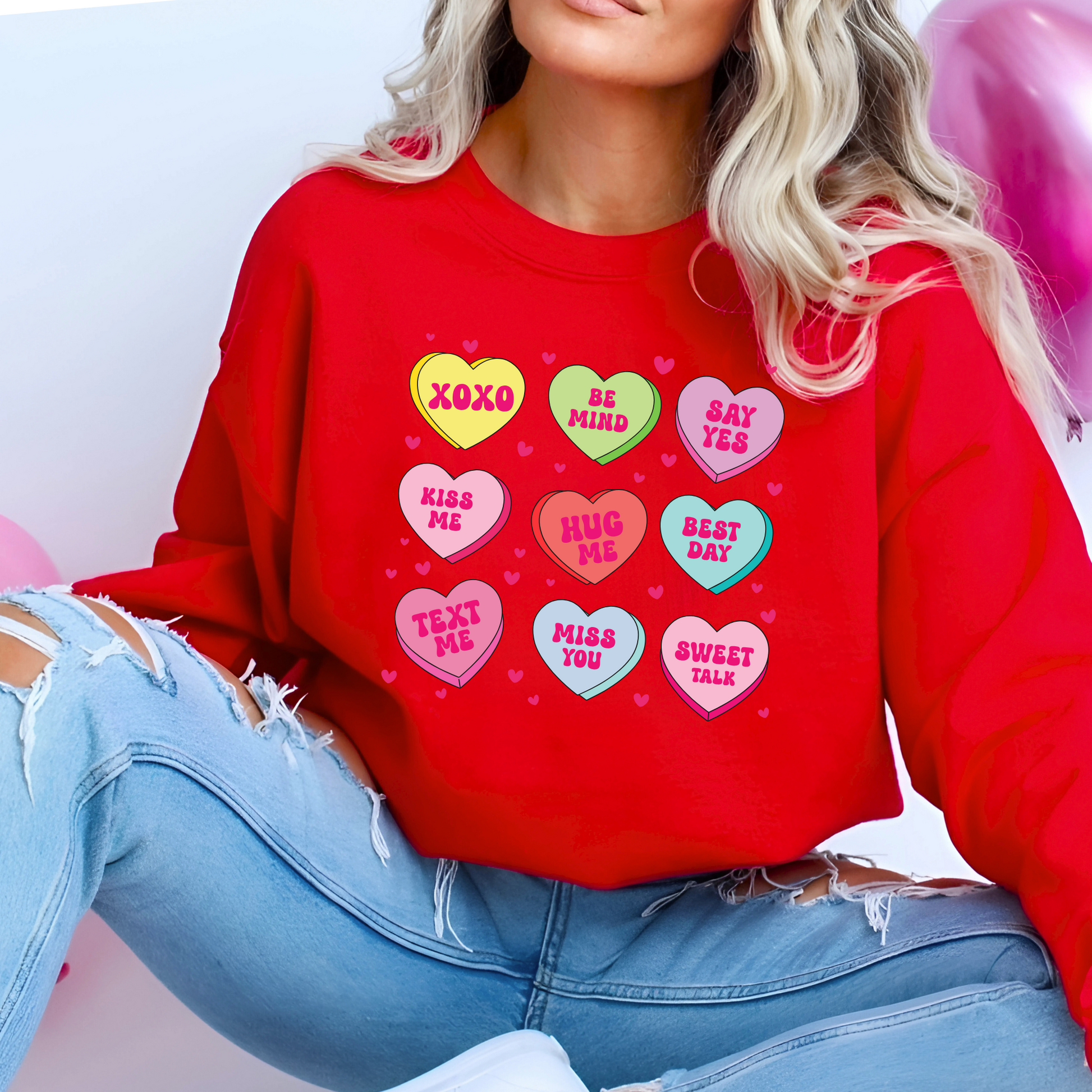 Woman wearing a red Valentine’s sweatshirt featuring colorful candy conversation hearts with messages like “XOXO,” “Be Mine,” “Say Yes,” “Kiss Me,” “Hug Me,” and more.