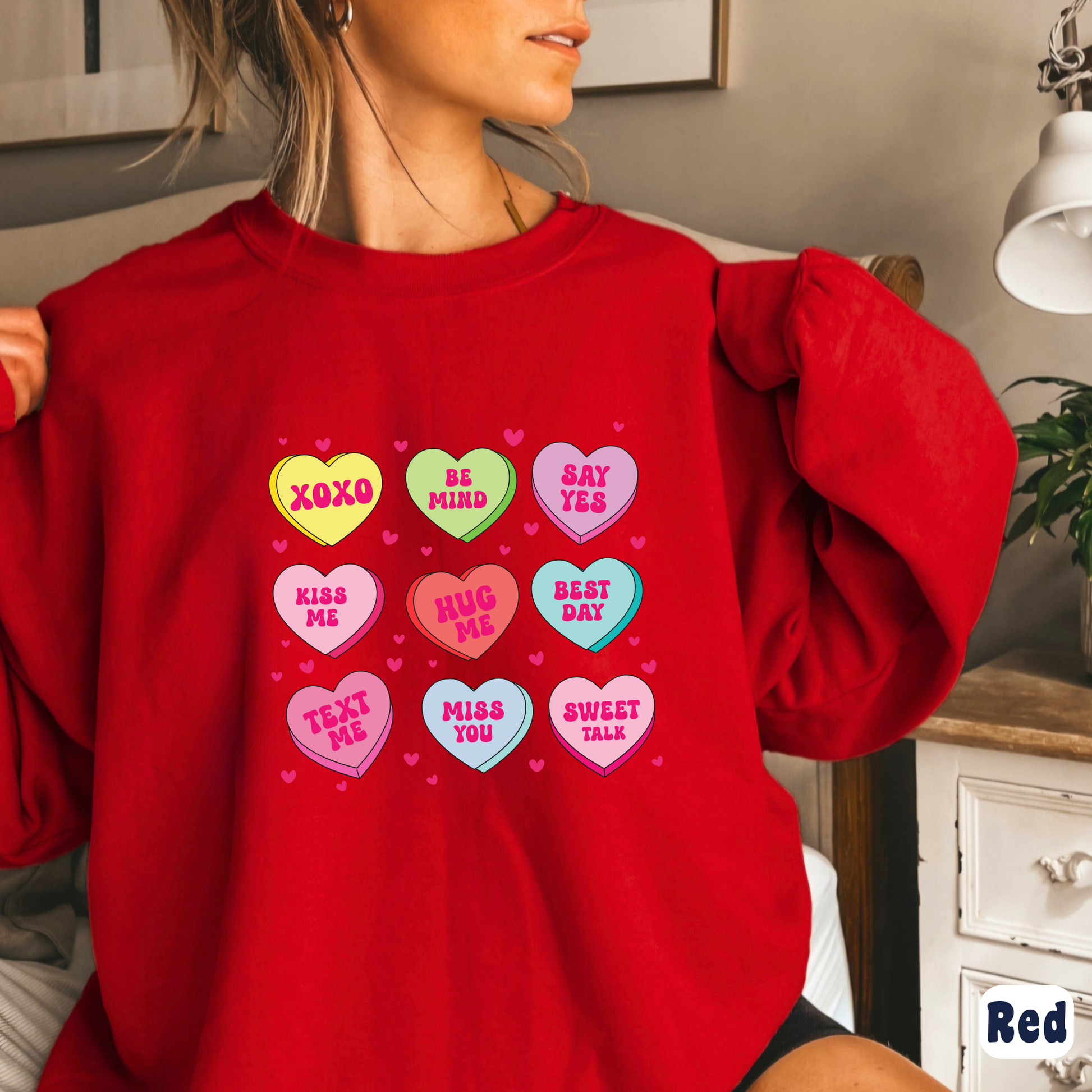 Woman wearing a red Valentine’s sweatshirt featuring colorful candy conversation hearts with messages like “XOXO,” “Be Mine,” “Say Yes,” “Kiss Me,” “Hug Me,” and more.