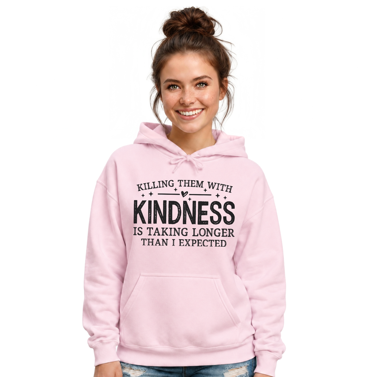Smiling woman modeling a light pink Killing Them With Kindness hoodie with jeans against a white background.