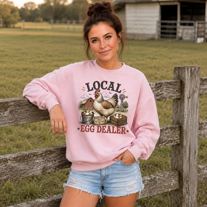 A woman wearing a light pink crewneck sweatshirt with a rustic 'LOCAL EGG DEALER' graphic featuring a chicken and eggs, leaning against a wooden fence in a farm setting.