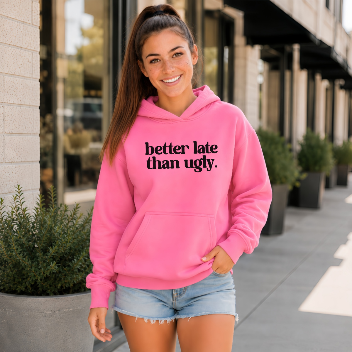 A woman wearing a bright pink pullover hoodie with the text 'better late than ugly.' printed in black across the chest, paired with light-wash denim shorts, standing outdoors on a sidewalk.