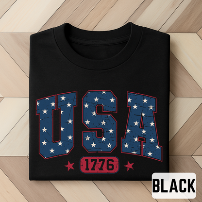Black folded T-shirt featuring bold USA 1776 graphic in distressed navy and red, patriotic flat lay style