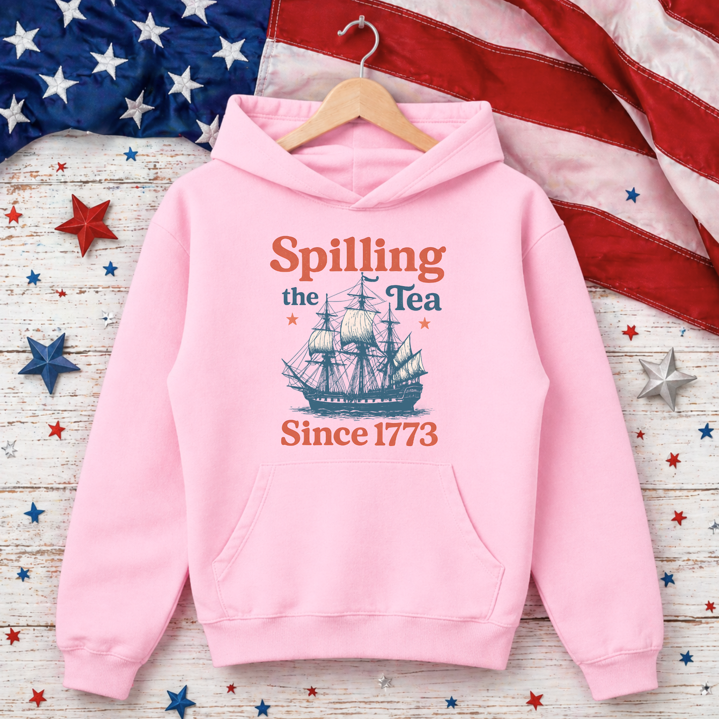 Pink hoodie with ship graphic and text 'Spilling the Tea Since 1773' on a wooden surface with American flag and stars.