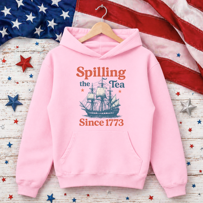 Pink hoodie with ship graphic and text 'Spilling the Tea Since 1773' on a wooden surface with American flag and stars.