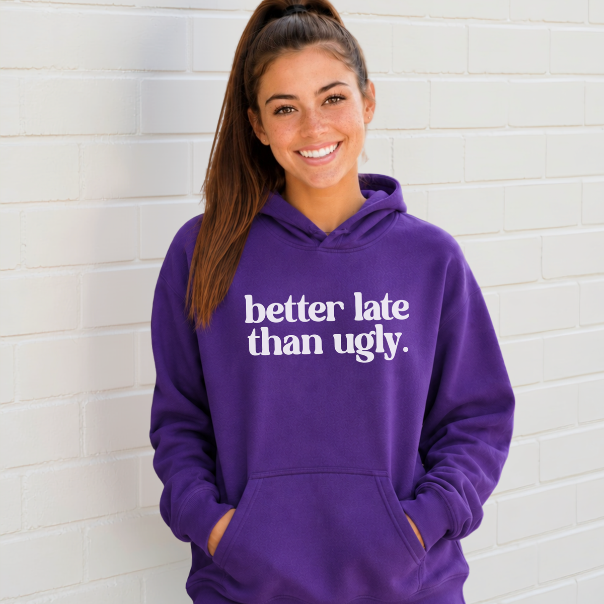 Woman wearing a purple hoodie  with white Better Late Than Ugly lettering against a white brick background.