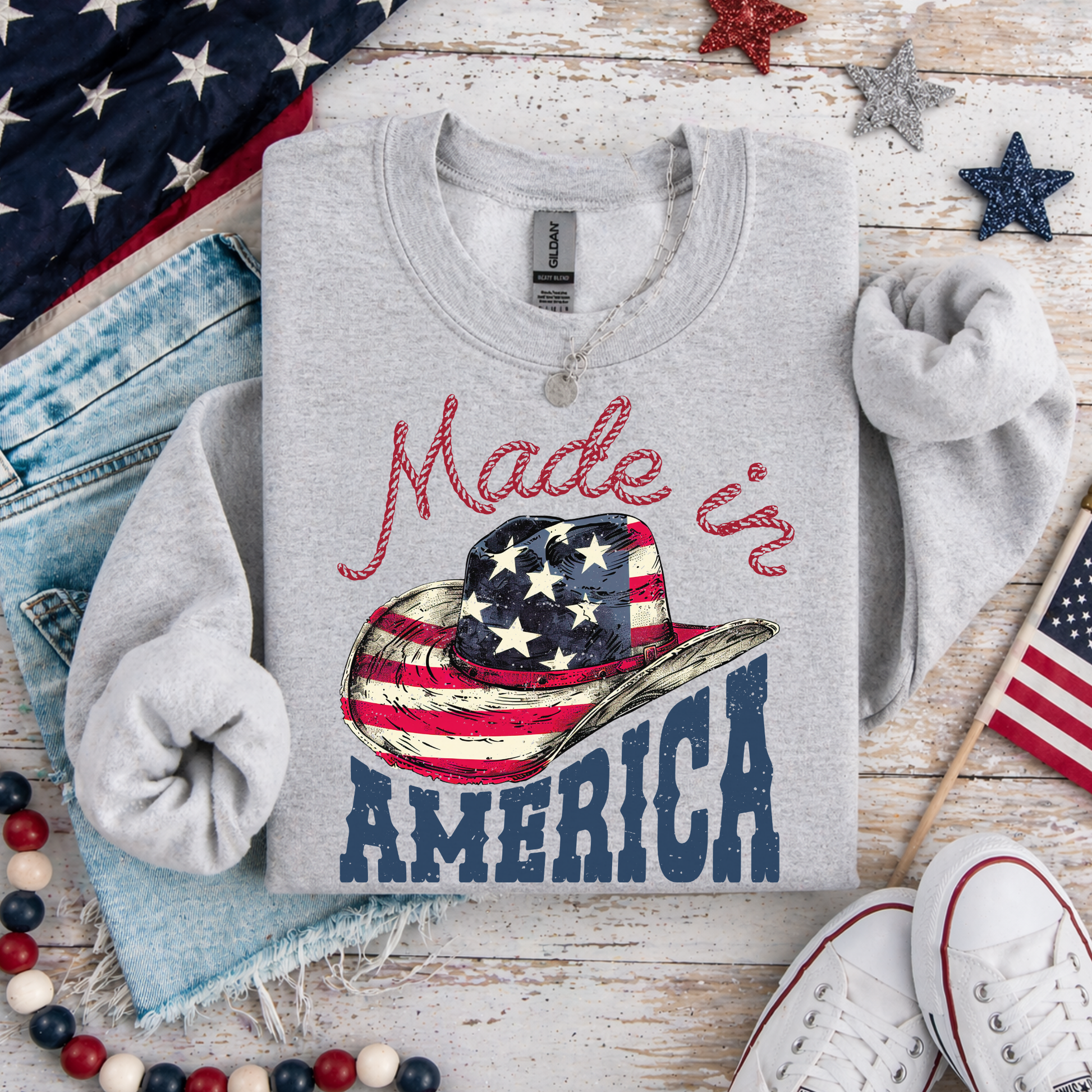 Gray sweatshirt with 'Made in America' design featuring a cowboy hat and American flag on a wooden surface with patriotic decor.