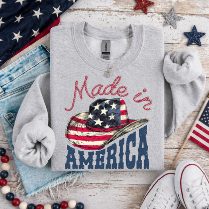 Gray sweatshirt with 'Made in America' design featuring a cowboy hat and American flag on a wooden surface with patriotic decor.