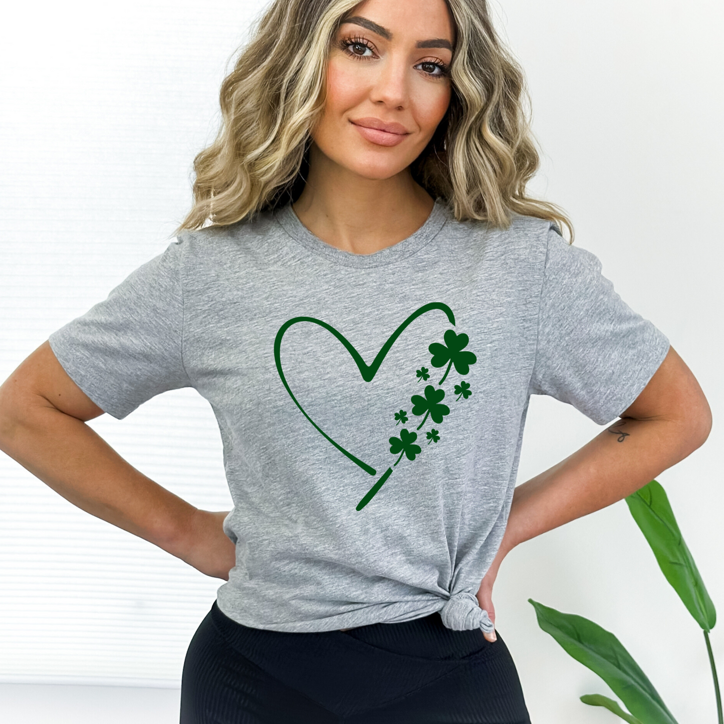A woman wearing a Gray T-shirt with a minimalist green heart and shamrock design, perfect for St. Patrick’s Day celebrations.