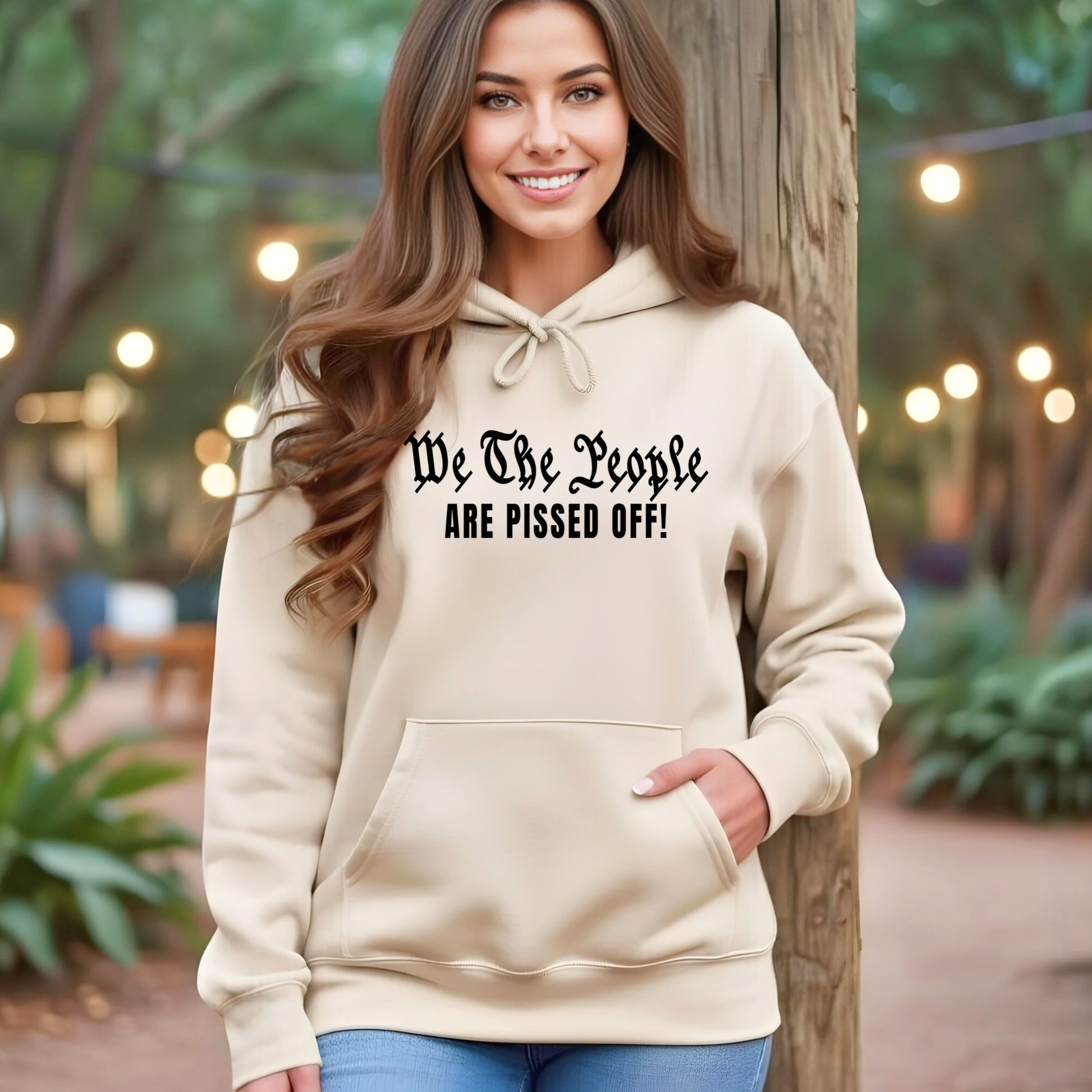 Woman wearing a beige hoodie with text 'We The People ARE PISSED OFF!' in an outdoor setting.