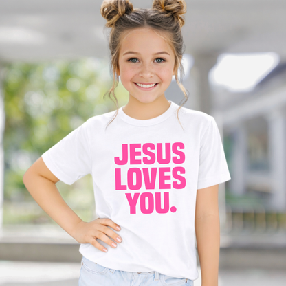 Young girl wearing a white t-shirt with 'Jesus Loves You' in pink text, standing outdoors.