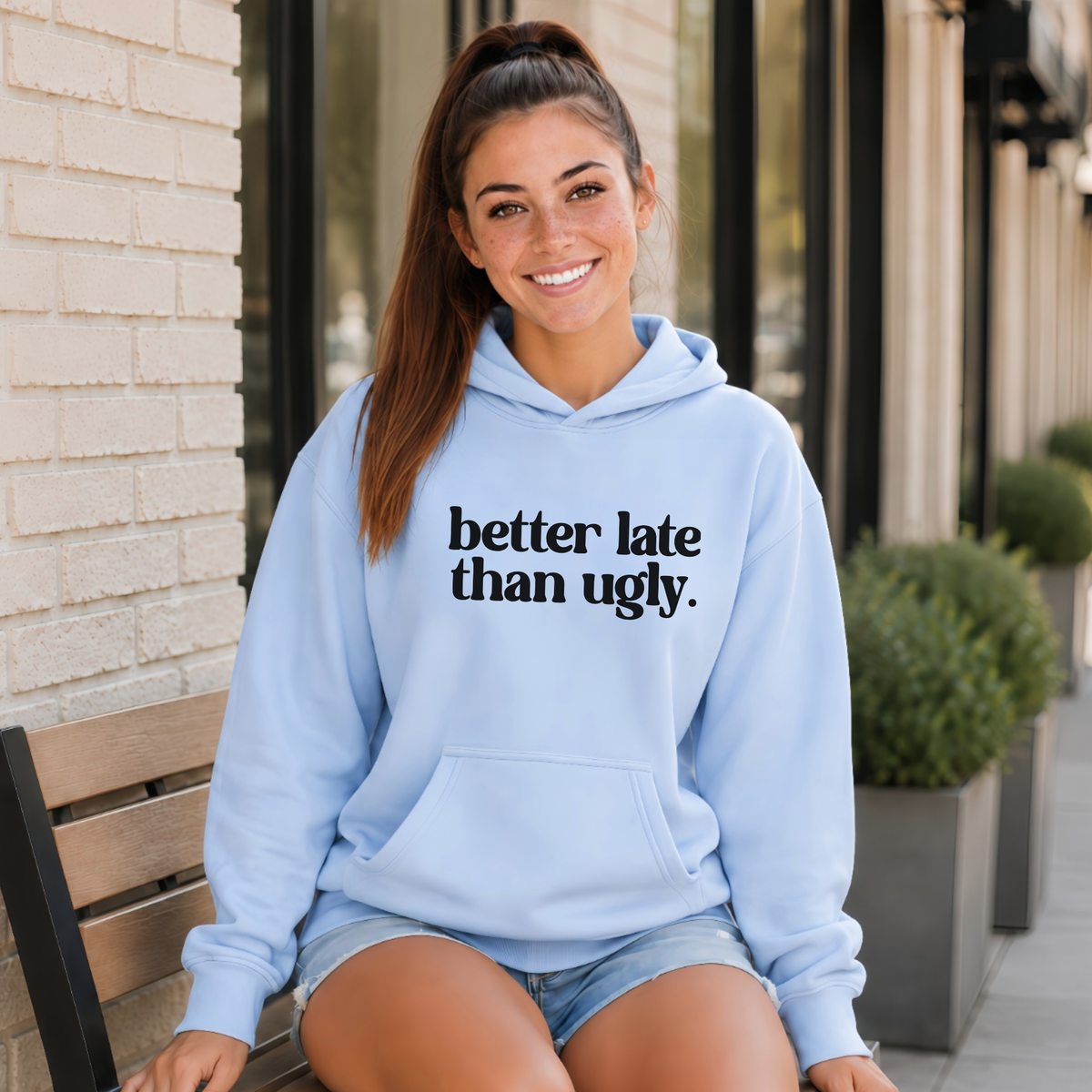 A woman sitting on a wooden bench outdoors wearing a light blue hoodie with the text 'better late than ugly.' printed in black across the chest, paired with denim shorts.