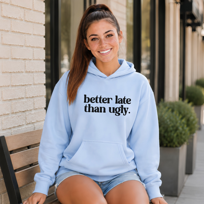 A woman sitting on a wooden bench outdoors wearing a light blue hoodie with the text 'better late than ugly.' printed in black across the chest, paired with denim shorts.