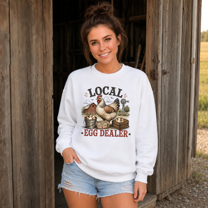A woman wearing a white crewneck sweatshirt with a rustic 'Local Egg Dealer' graphic featuring chickens and eggs, paired with denim shorts, standing in front of a weathered wooden barn door.
