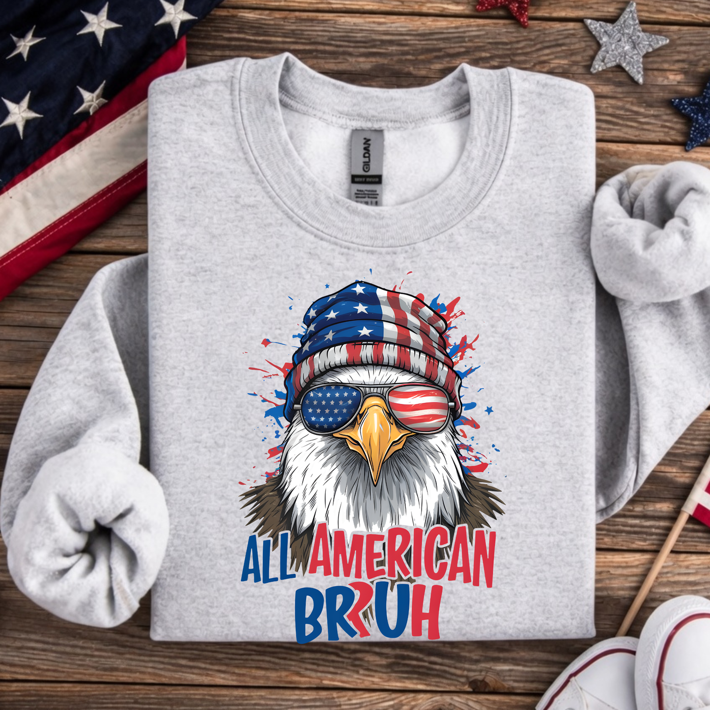 Gray sweatshirt with an eagle wearing a USA flag beanie and sunglasses, text 'All American Bruh' on a wooden surface with American flags.